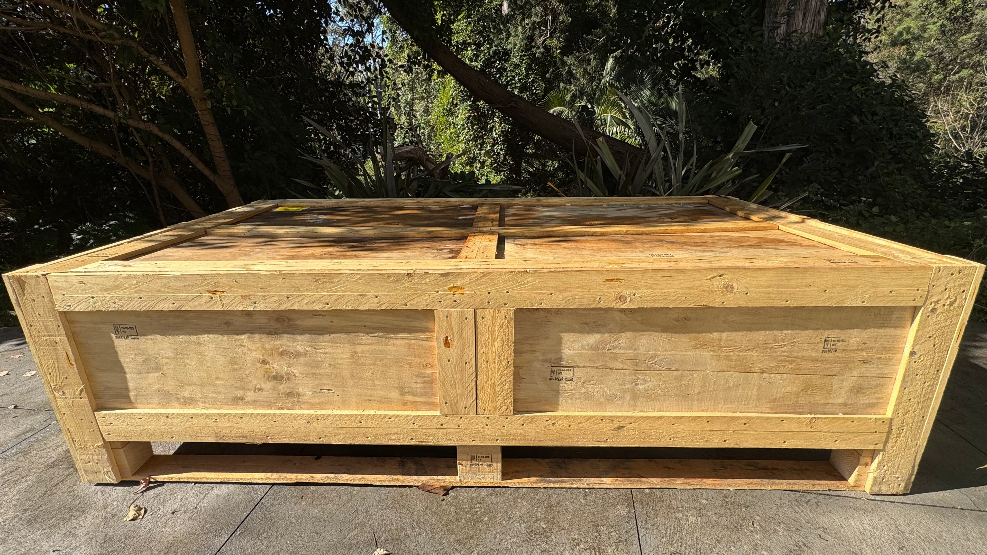 A large unfinished wooden storage chest or bench made of light natural wood, photographed outdoors on a concrete surface with trees and shrubs in the background.
